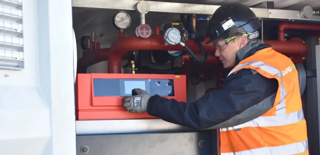 A Rapid Energy engineer wearing a black helmet, safety goggles, and an orange high-visibility vest operates the control panel of a temporary boiler unit.