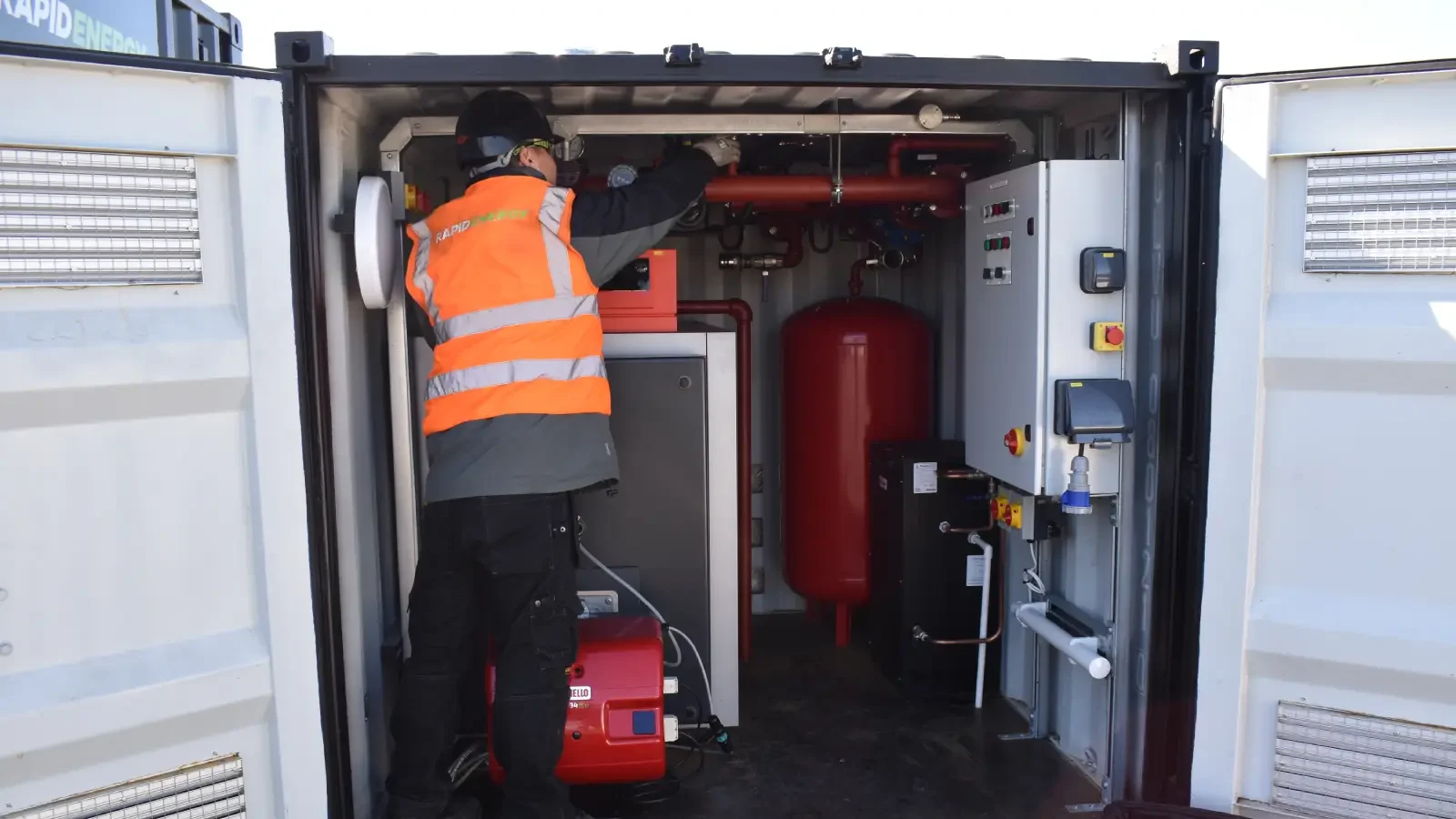 A Rapid Energy engineer wearing a black helmet, safety goggles, and an orange high-visibility vest operates the control panel of a temporary boiler unit.