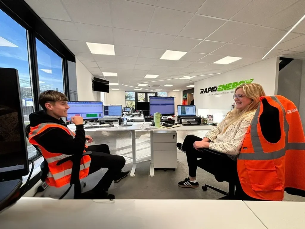 Rapid Energy Internal Sales Apprentice Harry is working in the office at his desk, chatting with a colleague.
